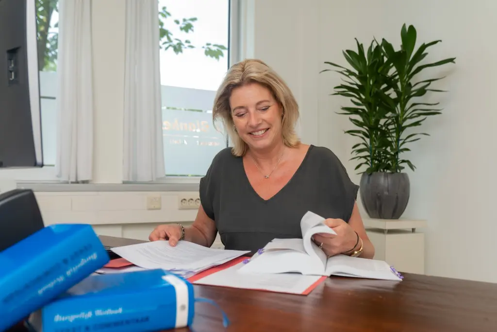 Vrouw zit aan bureau en bladert in wetboek en dossier